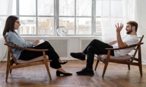 Psychiatrist and Person in Black Pants and Black Shoes Sitting on Brown Wooden Chairs While Talking